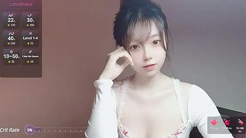 Media: Video of an Asian woman with light skin, medium-sized breasts, wearing a white top, sitting in a dimly lit room with a gray background.