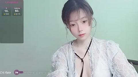 Media: A video of an Asian woman with fair skin and black hair tied back, wearing a white lace cardigan and a black necklace. The background is a light green wall with a digital thermometer.