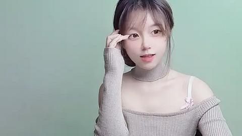 Media: Video of a young East Asian woman with light skin, straight black hair, and brown eyes. She wears a beige off-shoulder sweater with a white strap and small pink bow. Background is a plain, muted green.