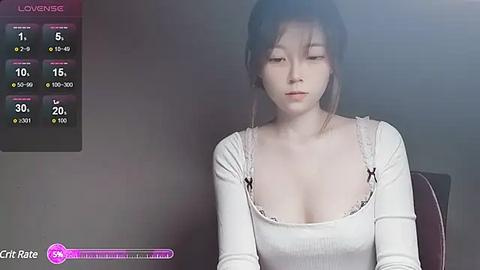 Media: Video of an Asian woman with fair skin, short brown hair, wearing a white, long-sleeved top with a low neckline, sitting indoors. The background is dimly lit with a blurred view.