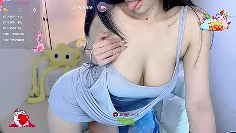 Media: A video of an Asian woman with long black hair, wearing a tight, light blue tank top, exposing ample cleavage. She is in a room with a yellow SpongeBob SquarePants plush toy.