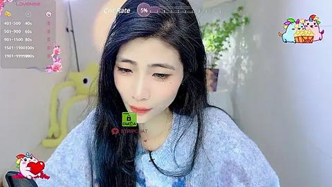 Media: Video of a young East Asian woman with long black hair, fair skin, and a sad expression, wearing a blue sweater. Background features a white room with a yellow chair, potted plants, and a computer screen displaying social media chat.