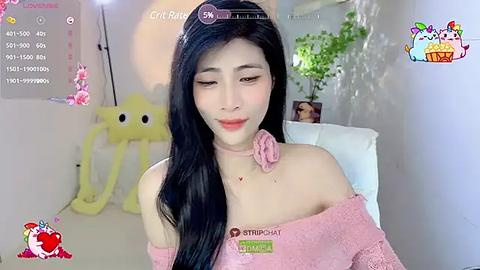 Media: Video of a young Asian woman with long black hair, wearing a pink off-shoulder sweater, sitting on a white chair. Background features a yellow plush toy, green plants, and a partially visible wall.