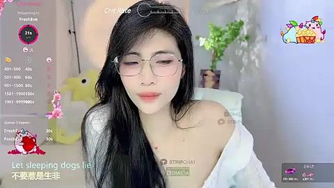 Media: Video of an Asian woman with long black hair, wearing glasses and a white off-shoulder top, in a cozy room with a pink flamingo toy and potted plants, captioned \"Let sleeping dogs lie.\