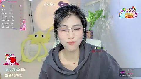 Media: Video of a young Asian woman with glasses, fair skin, and black hair, wearing a dark hoodie, in a bright room with a yellow toy and green plants.