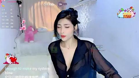 Media: A video of an East Asian woman with fair skin, black hair styled in a bun, wearing a sheer black blouse, seated indoors. The room has a white theme with a pink bunny and a calendar.