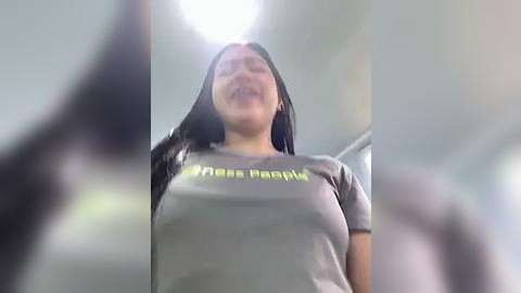 Media: Video of a young woman with long black hair, wearing a gray t-shirt with \"My Name is\" written in yellow on the chest, smiling broadly, taken from a low angle.