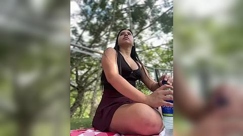 Media: A video of a woman with long black hair, wearing a black tank top and shorts, meditating outdoors on a green grassy area, surrounded by trees and a picnic blanket.