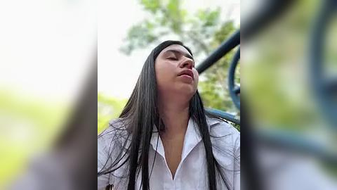 Media: Video of a young woman with long, straight black hair, wearing a white button-up shirt, standing outside with blurred green foliage and metal railing in the background, eyes closed and mouth slightly open.