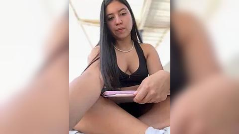 Media: Video of a Latina woman with long black hair, wearing a black bra and shorts, sitting on a bed with legs spread.