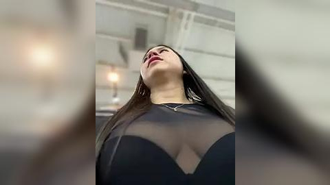 Media: A video captures a woman with long dark hair, wearing a black sheer top that reveals her ample cleavage, standing in a modern, industrial-style room with white walls and metal beams.