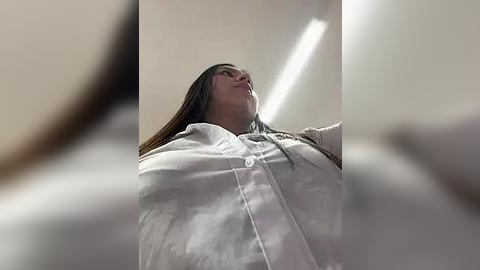 Media: Video of a woman in a white lab coat looking upwards, blurred background, fluorescent light overhead, focused on her face.