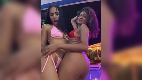 Media: Video of two women in bikinis, one pink, one red, embracing passionately in a dimly-lit nightclub with blurred backgrounds.