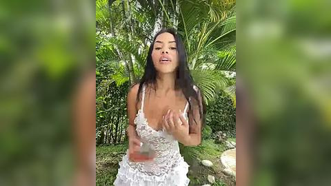 Media: Video of a young woman with long dark hair, wearing a white lace dress, standing in a lush green garden with palm trees, smiling and covering her chest.