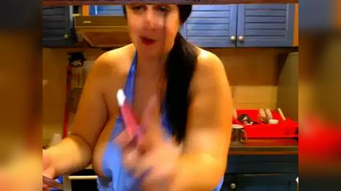 Media: A video of a fair-skinned, curvaceous woman with long black hair, wearing a blue apron, scrubbing a countertop in a kitchen with wooden cabinets. The image is slightly blurred.