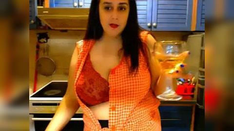 Media: A video of a plus-size woman with long black hair, red lipstick, and a red lace bra, standing in a modern kitchen with beige walls, blue cabinets, and a stove.