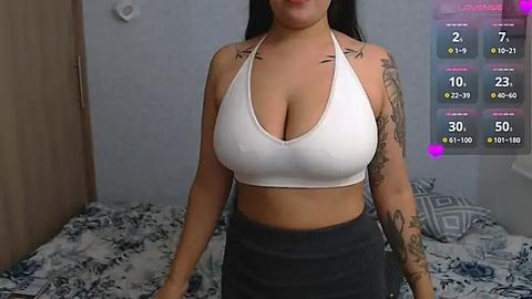 Media: Video of a woman with long black hair, wearing a white halterneck sports bra and high-waisted gray shorts, standing in a bedroom with blue and white bedding.