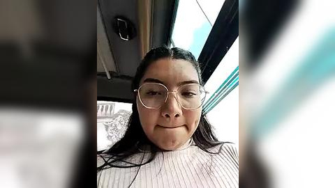 Media: A video of a young woman with long dark hair and glasses, wearing a ribbed white turtleneck, smiling inside a car with a blurred exterior.