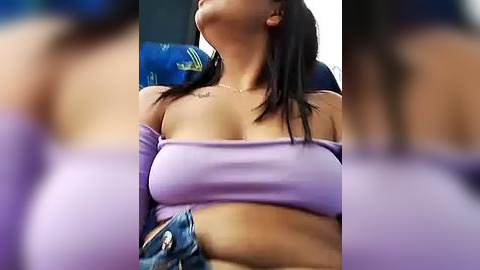 Media: Video of a woman with medium brown skin, dark hair, and a tattoo on her collarbone, wearing a purple off-shoulder top, sitting indoors with a blurred background.