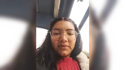 Media: Video of a woman with long, curly hair, wearing glasses and a red sweater, sitting in a car. Her expression is neutral.