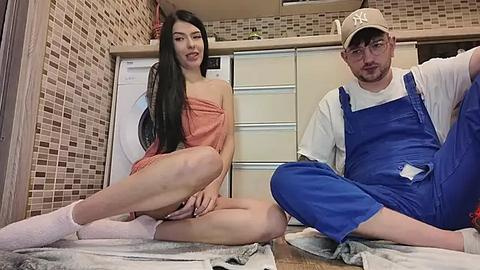 Media: Video of an East Asian woman in a strapless peach dress and white socks, and a bearded Caucasian man in blue overalls and a Yankees cap, sitting on a tiled floor in a kitchen during renovation.