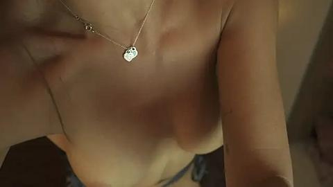 Media: A close-up video of a topless woman with light skin, showing her bare breasts, wearing a delicate silver heart necklace, and a blurred background.