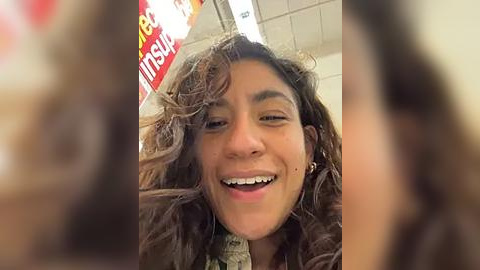 Media: A blurry video captures a joyful, smiling woman with curly hair and a green shirt, standing in a brightly lit supermarket with colorful signage in the background.