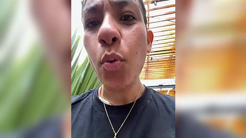 Media: Video of a young woman with light brown skin, short hair, and dark eyes, puckering her lips in a playful manner. She wears a black t-shirt and a gold necklace. Background includes green palm leaves and wooden slats.