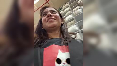 Media: A video of a woman with long, dark hair wearing a gray t-shirt featuring a cartoon cat, grinning widely, standing in a laundry room with neatly stacked white towels.