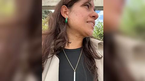 Media: Video of a woman with long brown hair, wearing a black t-shirt, beige jacket, and green earrings, smiling outdoors with blurred background.