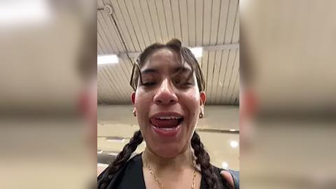 Media: A video of a young woman with long, braided hair, light skin, and a black top, smiling broadly and sticking out her tongue. The background is an indoor gymnasium with a white ceiling and fluorescent lights.