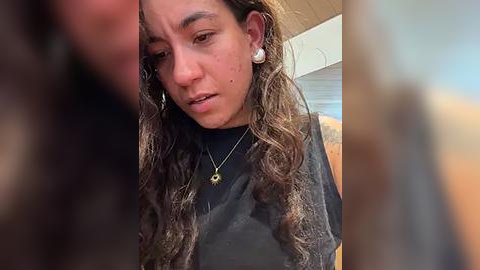 Media: A candid video of a young woman with long, wavy brown hair, wearing a black shirt, and a necklace. She appears to be indoors, looking downward with a somber expression.