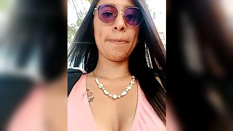 Media: Video of a woman with long black hair, wearing pink sunglasses, a pink top, and a pearl necklace, taken from a side angle, showing her facial scars and tattoos.