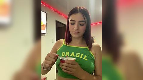 Media: Video of a young woman with straight, shoulder-length auburn hair, wearing a green \"Brazil\" crop top, stirring a drink with a spoon. Background features red LED lights and a blurred person.