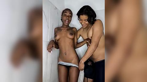 Media: Video of two topless women in a shower, one with small breasts, the other with medium breasts. Both wear only panties, one pulling down the other's underwear. Background features white tiled walls.