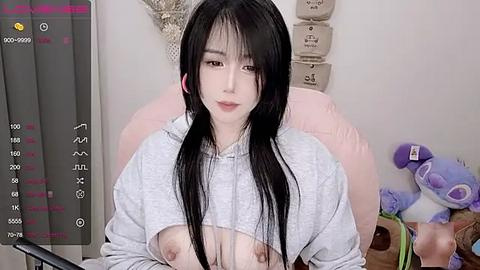 Media: Video of an East Asian woman with long black hair, wearing a gray hoodie lifted to reveal her bare breasts. Background includes plush toys and a recording overlay.