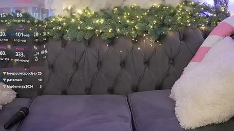 Media: Video of a cozy living room with a plush gray couch, covered in white fur, adorned with a decorative green garland and twinkling lights. The background features a TV displaying social media stats.