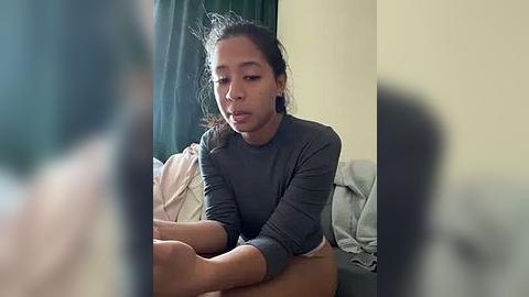 Media: Video of a young woman with medium brown skin, dark hair tied back, wearing a grey long-sleeved top, sitting on a bed, looking thoughtful, in a softly lit room with green curtains and beige walls.