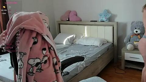 Media: Video of a cozy, minimalist bedroom with a white bed, gray sheets, plush toys, and a pink blanket featuring cartoon dogs.