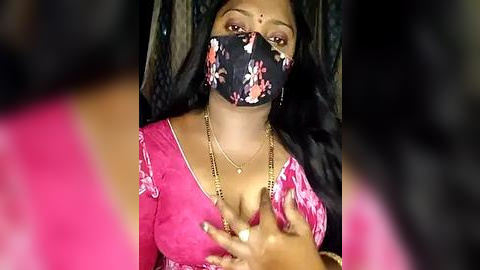 Media: Video of a woman with dark skin and long black hair, wearing a pink floral dress, black floral face mask, and gold jewelry, holding her chest, with blurred background.