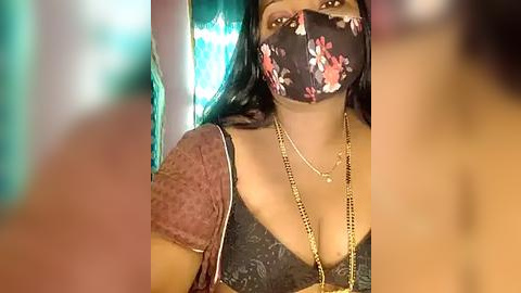 Media: Video of a woman with medium-dark skin tone, wearing a black floral mask, black lace top, gold necklace, and teal background, possibly indoors.