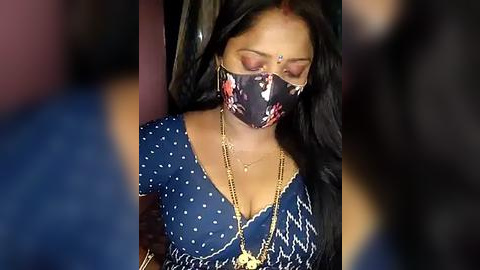 Media: Video of a South Asian woman with medium brown skin, long black hair, wearing a blue polka-dot blouse, a black floral mask, and gold necklaces. Background is dark and blurry, focusing on her.