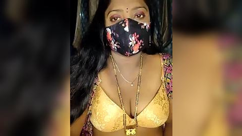 Media: Video of a South Asian woman with dark skin and long black hair, wearing a floral face mask, yellow lace bra, and gold necklace, indoors.