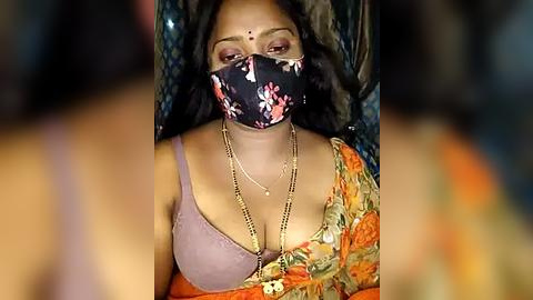 Media: Video of a South Asian woman with medium skin tone, wearing a black floral mask, lavender bra, orange sari, and gold necklace, seated against a dark, blurry background.