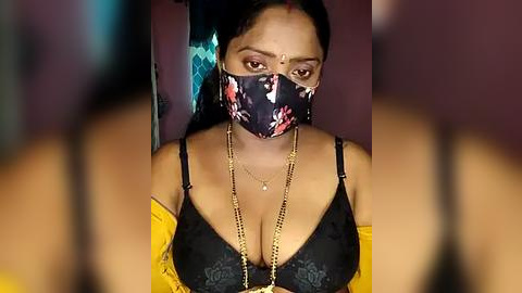 Media: Video of a South Asian woman with medium brown skin, wearing a black floral mask, black lace bra, and yellow blouse, with a necklace, in a dimly lit room.