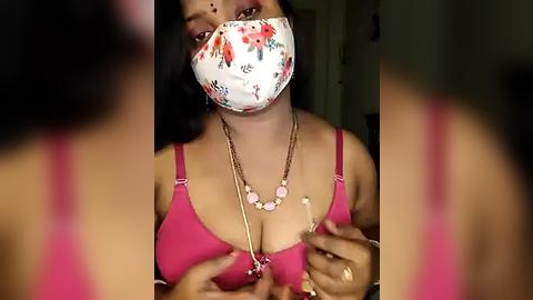 Media: Video of a woman in a pink bra, wearing a floral face mask, gold necklace, and holding her breasts.