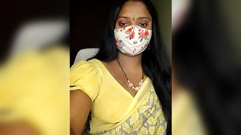 Media: Video of an Indian woman wearing a floral face mask and yellow saree with a floral pattern, seated indoors against a dark background.