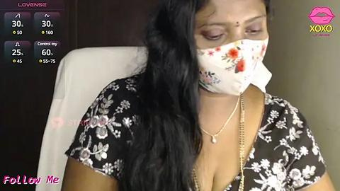 Media: Video of a South Asian woman with long black hair, wearing a floral-patterned mask, black blouse, and gold necklace, seated indoors.