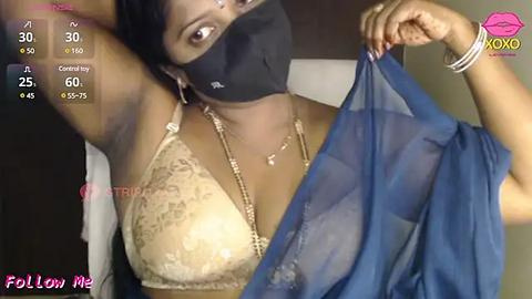 Media: A video of a dark-skinned woman in a lace bra and blue veil, wearing a black face mask, holding the veil up. Background is blurred.