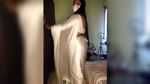 Media: A video of a woman with long black hair, wearing a white sari and mask, standing in a dimly lit room with green walls and a bed.